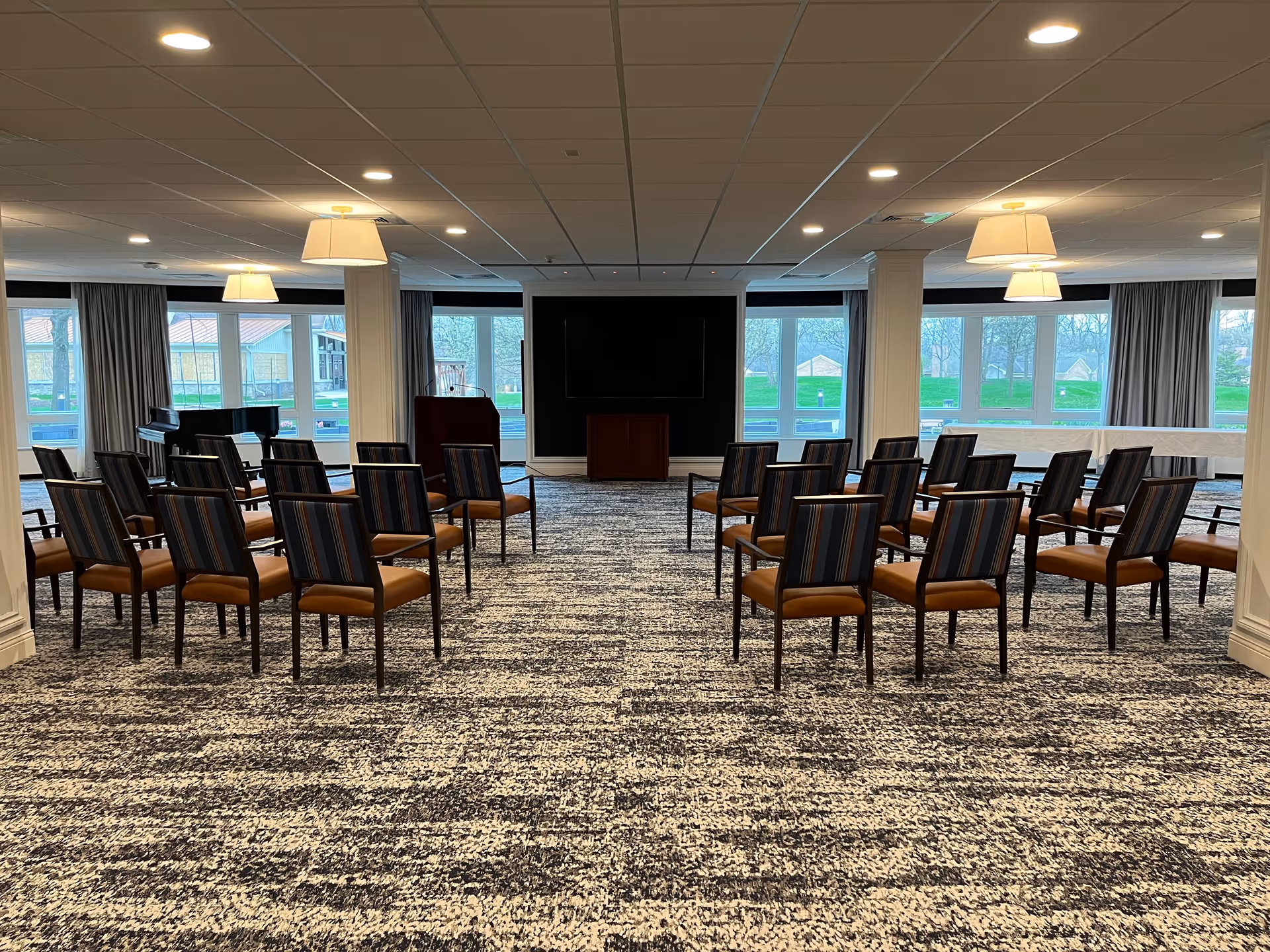 A spacious meeting or event room with rows of chairs arranged facing a podium and a large flat screen on the wall. The room has large windows with views of greenery outside, carpeted floor, and ceiling lights with hanging lampshades.