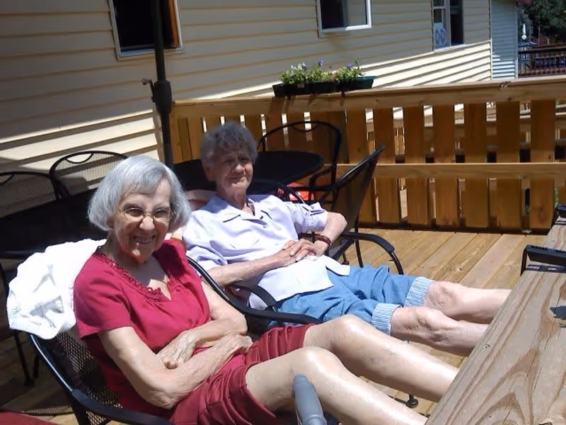 Two elderly women sitting and relaxing on black metal chairs on a wooden deck outside a building with beige siding. One woman is wearing a red outfit and glasses, smiling at the camera, while the other woman is wearing a light purple shirt and blue shorts, sitting back with her hands folded.