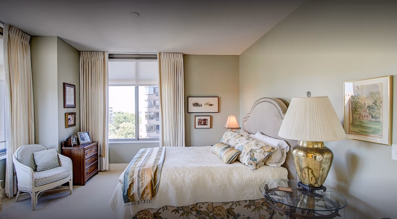 Sunlit, traditionally furnished bedroom with a bed, bedside lamps, an armchair, a dresser, and a large window overlooking trees and a nearby building.