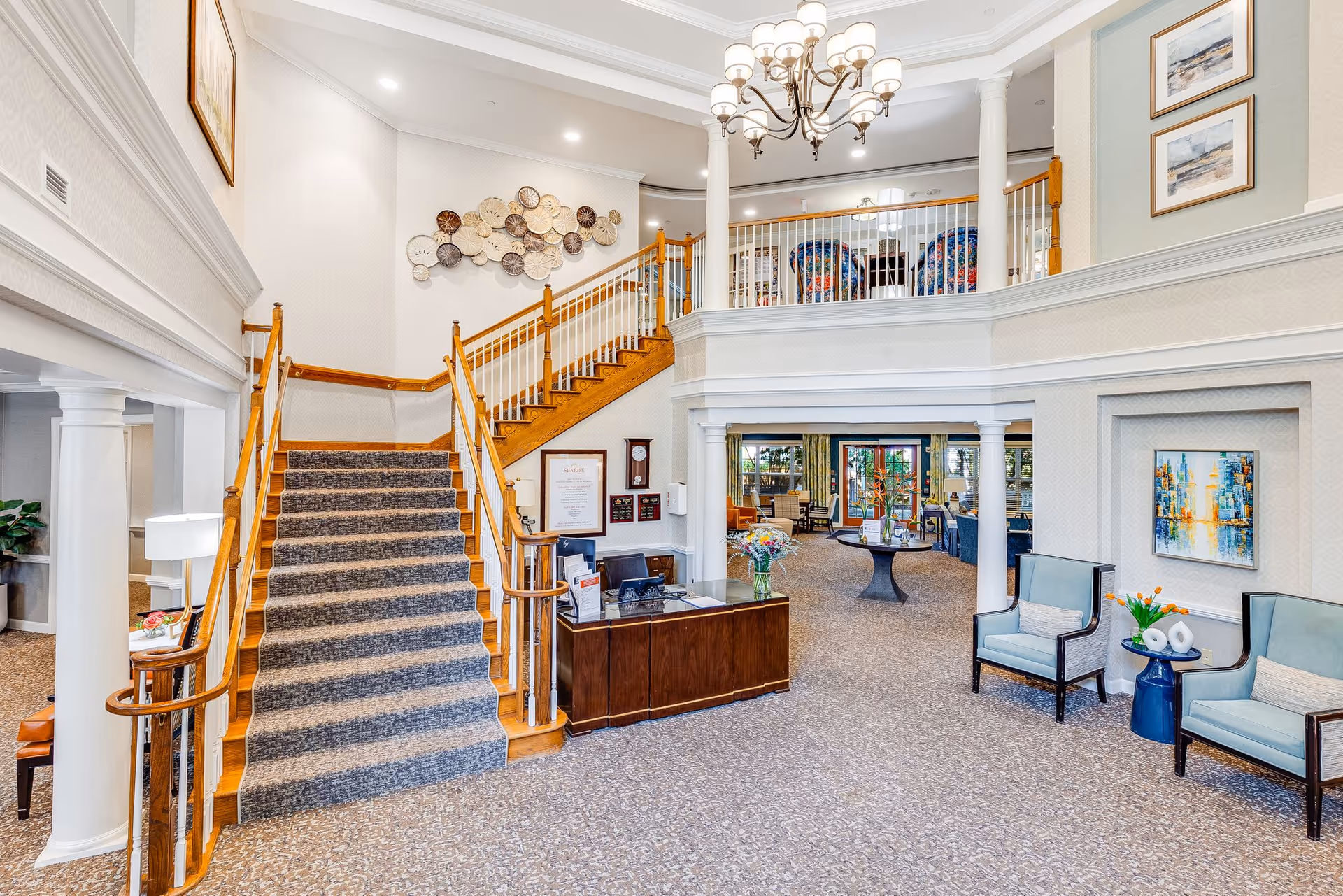 Bright two-story lobby with a central carpeted staircase, reception desk, seating areas, and decorative artwork.