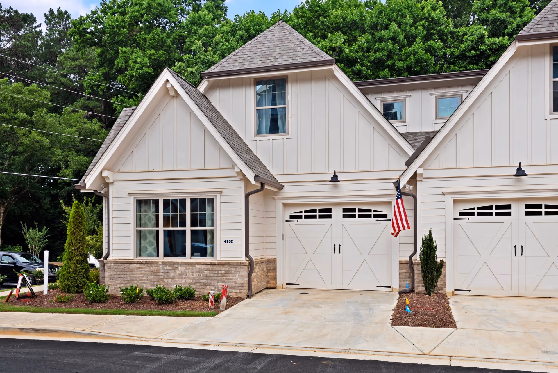 Front exterior of a light-colored townhouse with two garage doors, a window, landscaping, and an American flag.