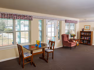 A cozy interior room with large windows letting in natural light. There is a small wooden table with two chairs, a Connect Four game on the table, a red upholstered armchair with a side table holding a lamp and a framed picture, and a bookshelf filled with books. The room has beige walls, a carpeted floor, and floral valances above the windows.