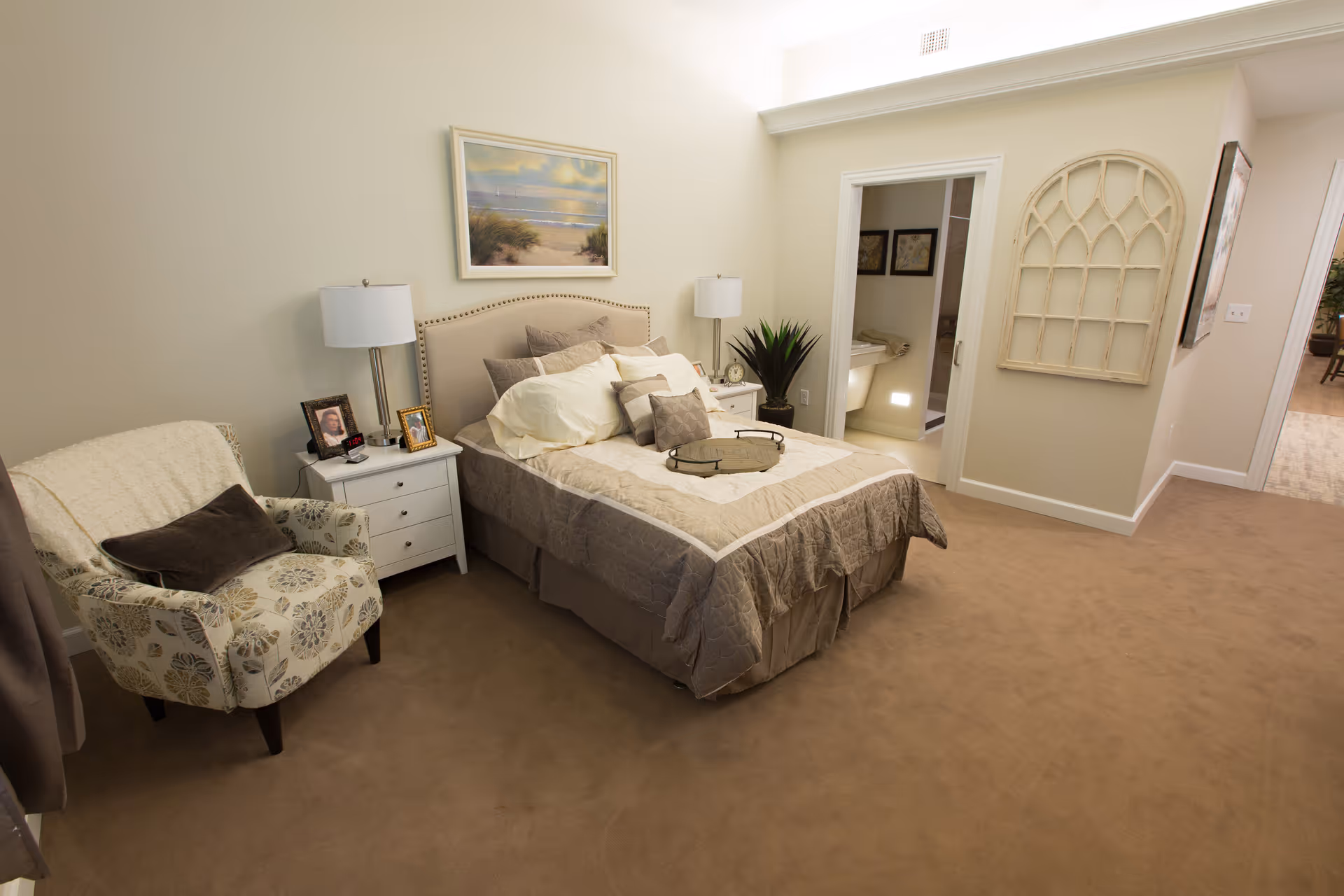 A cozy bedroom with a neatly made bed featuring multiple pillows and a tray on top. There is a patterned armchair with a dark cushion and a light throw blanket to the left of the bed. Two white nightstands flank the bed, each with a lamp and framed photos. A framed beach scene hangs on the wall above the headboard. To the right, an open door reveals a bathroom with a sink and mirror. The room has beige carpeting and light-colored walls with decorative wall art near the bathroom door.