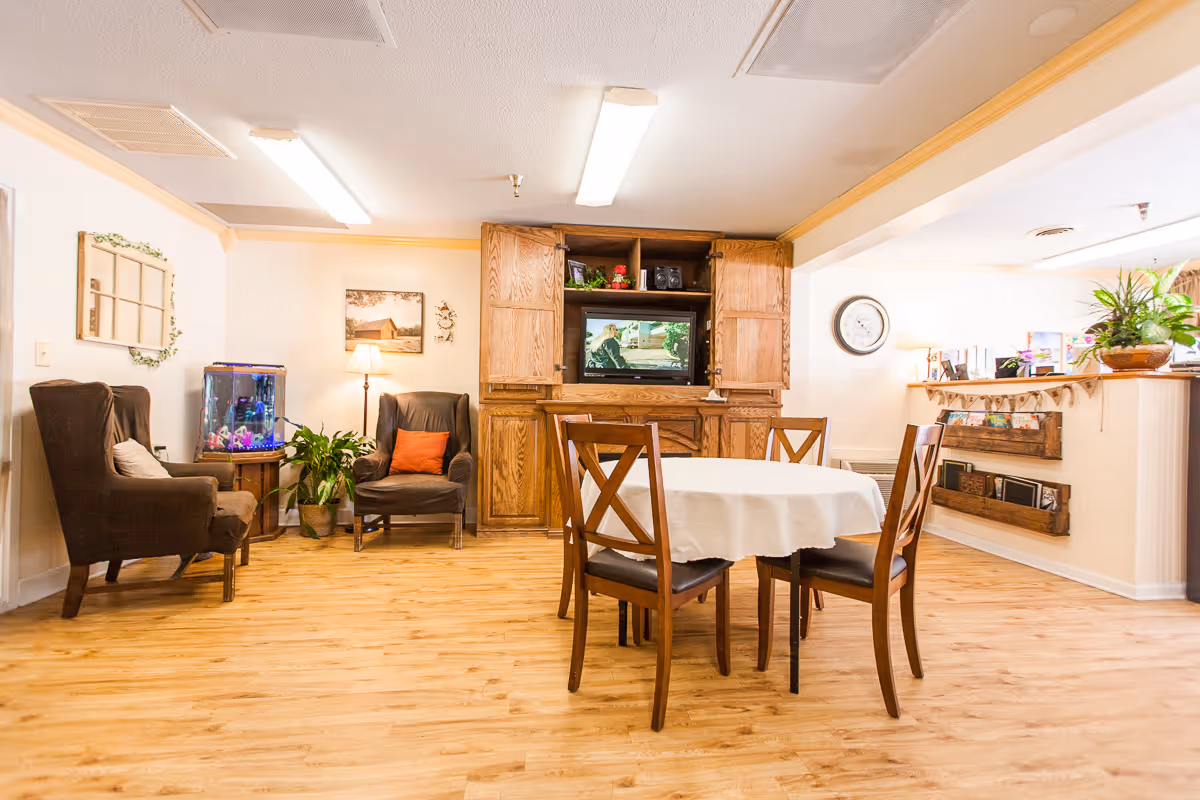 A cozy common area in a senior living facility featuring a round table with four wooden chairs covered with a white tablecloth in the center. To the left, there are two brown armchairs with cushions, a small table with a fish tank, and a potted plant. In the background, a wooden entertainment center holds a television and decorative items. The room has light-colored walls, wooden flooring, and ceiling lights. On the right side, there is a counter with plants and magazines displayed underneath.