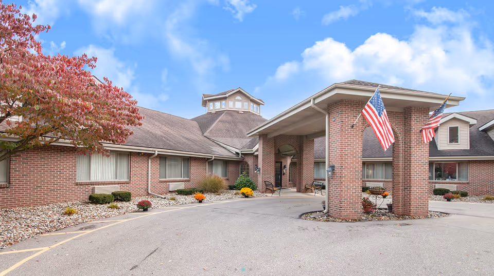 Exterior view of Brentwood at Elkhart Assisted Living facility showing a brick building with a covered entrance supported by brick pillars, two American flags, a tree with red leaves, and landscaping with rocks and flowers under a partly cloudy blue sky.