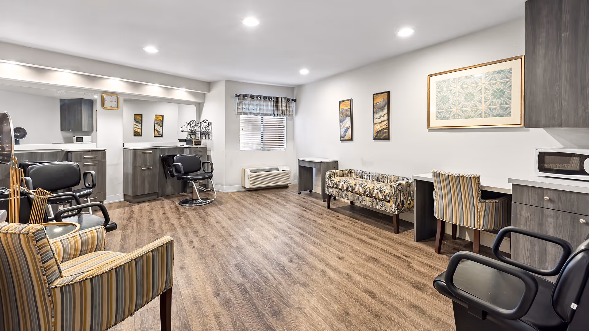A spacious, well-lit interior room with wood flooring, featuring salon-style black chairs, a striped armchair, a patterned bench, desks with chairs, and cabinets with a microwave. The walls are decorated with framed artwork and a window with blinds and a valance.