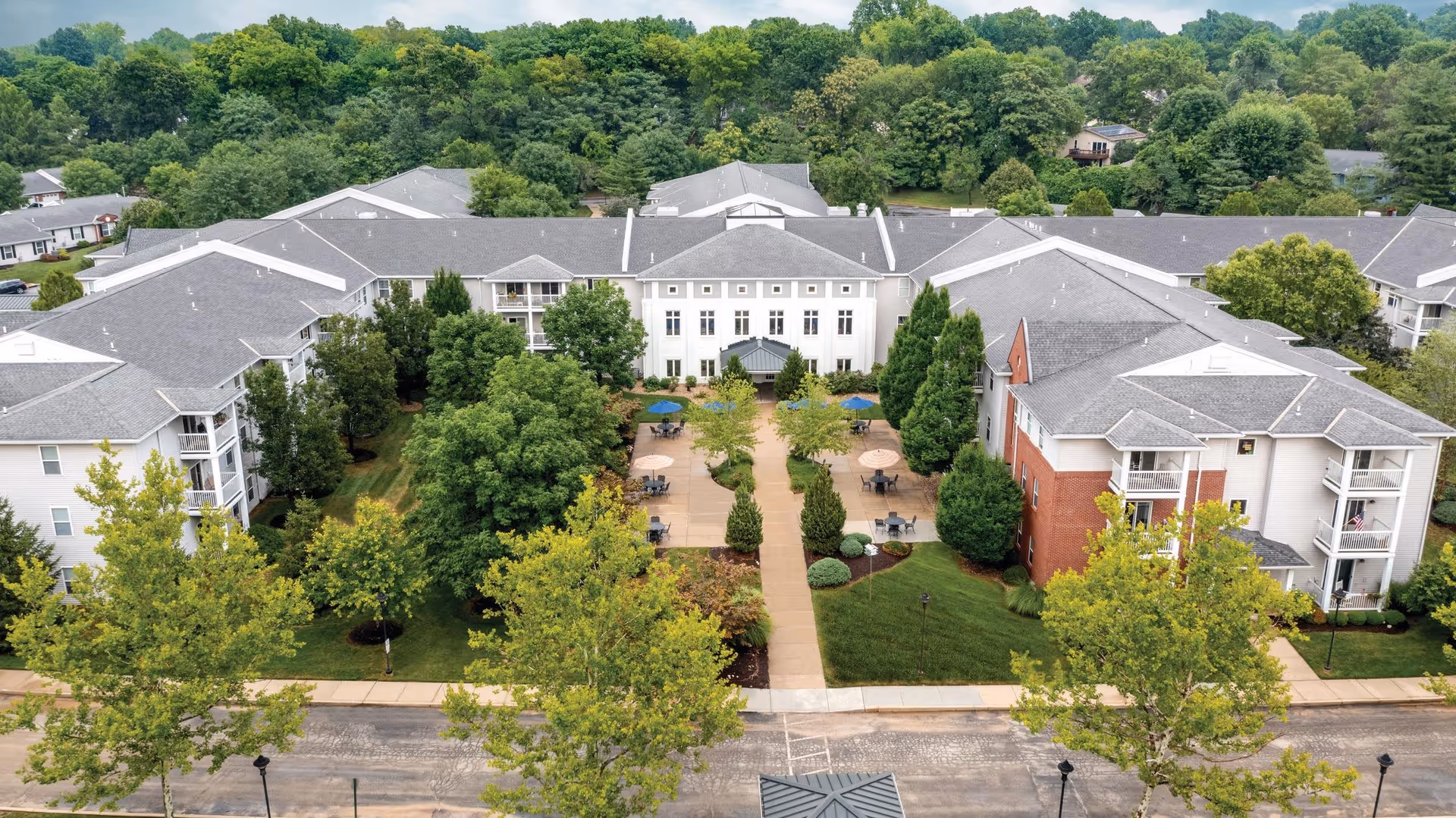 Aerial view of The Emerson at St. Peters senior living facility showing a large U-shaped building with gray roofs surrounded by lush green trees and landscaped gardens. The central courtyard features paved walkways, outdoor tables with umbrellas, and well-maintained shrubbery. The background includes dense forested areas.