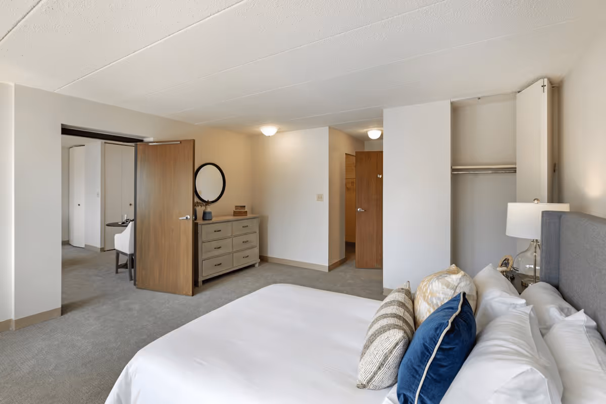 A spacious senior living bedroom with a neatly made bed featuring multiple pillows, a bedside table with a lamp, an open closet, a dresser with a round mirror above it, and an open door leading to another room with a chair and table visible.