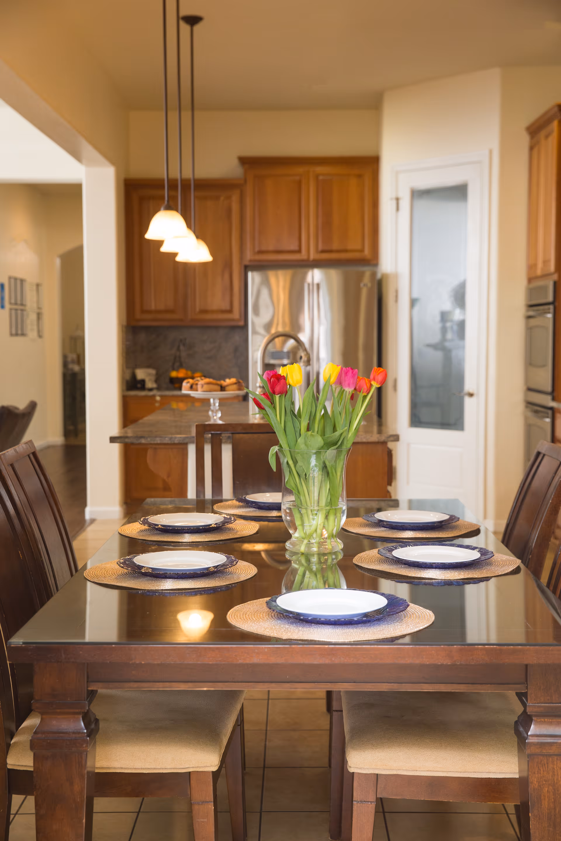 A dining table set for six with plates and a vase of tulips in an open dining area leading to a kitchen.