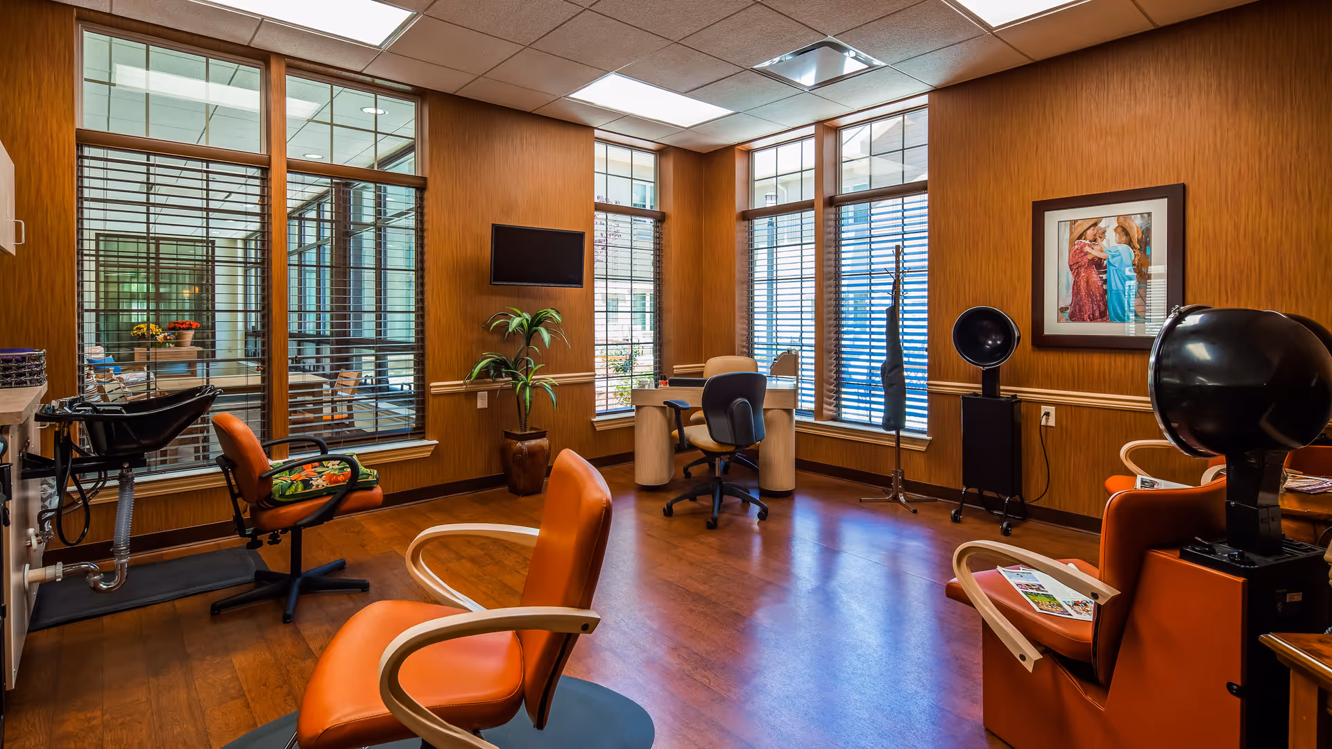 Interior view of a salon area with wooden floors and walls, featuring orange salon chairs, hair washing station, hair dryers, a desk with office chairs, large windows with blinds, a potted plant, and a framed picture on the wall.