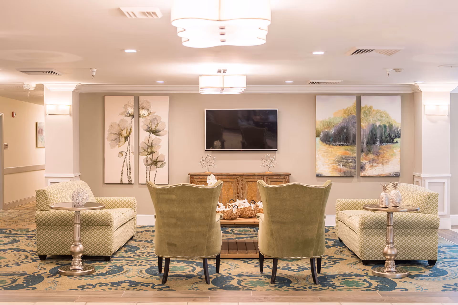 A cozy living room area in Royal Oaks Assisted Living Center featuring two green upholstered chairs and two patterned sofas arranged around a wooden coffee table. The room has a patterned carpet, a flat-screen TV mounted on the wall, and decorative paintings on either side of the TV. Soft ceiling lights illuminate the space.