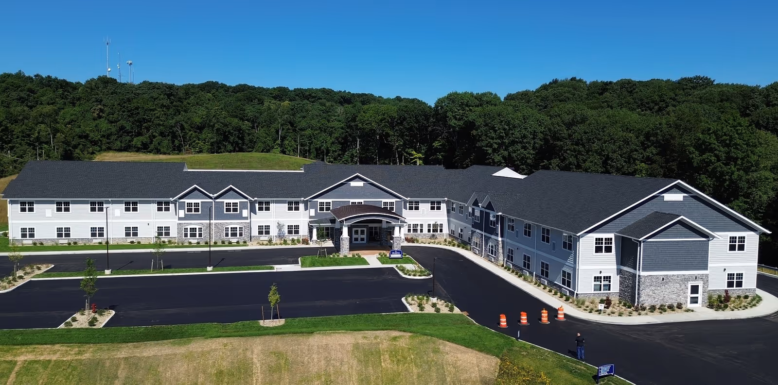 A large two-story senior living facility building with gray and white exterior walls, a dark roof, and a covered entrance. The building is surrounded by a paved parking lot with a few small trees and landscaped areas. Behind the building is a dense forest under a clear blue sky.