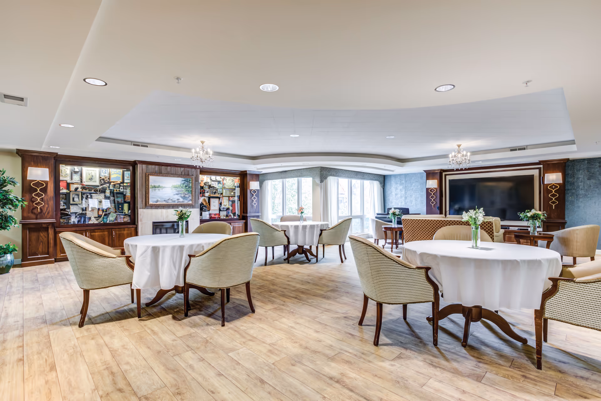A spacious, well-lit common area with round tables covered in white tablecloths, surrounded by cushioned chairs. The room features wooden flooring, large windows letting in natural light, decorative chandeliers, and a large flat-screen TV mounted on the wall. There are also display cabinets with framed pictures and decorative items.