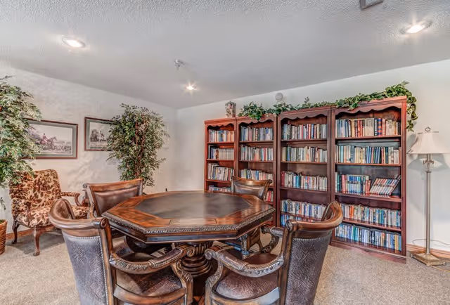 Comfortable reading room with a wooden card table surrounded by leather chairs and bookshelves filled with books.