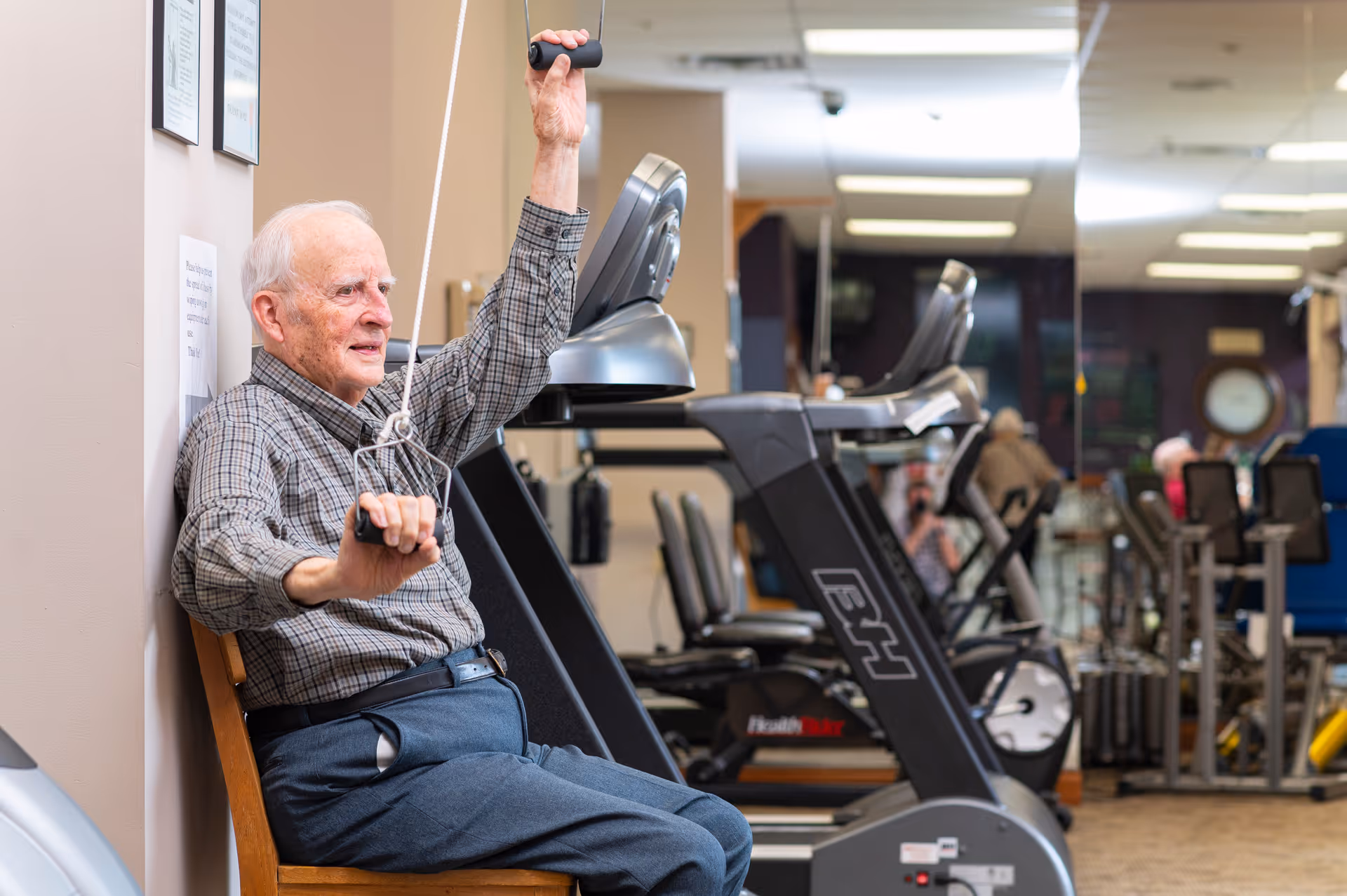 An elderly man sitting on a chair in a fitness room, using a pulley exercise machine. Behind him are treadmills and other gym equipment, with mirrors on the wall reflecting the room.