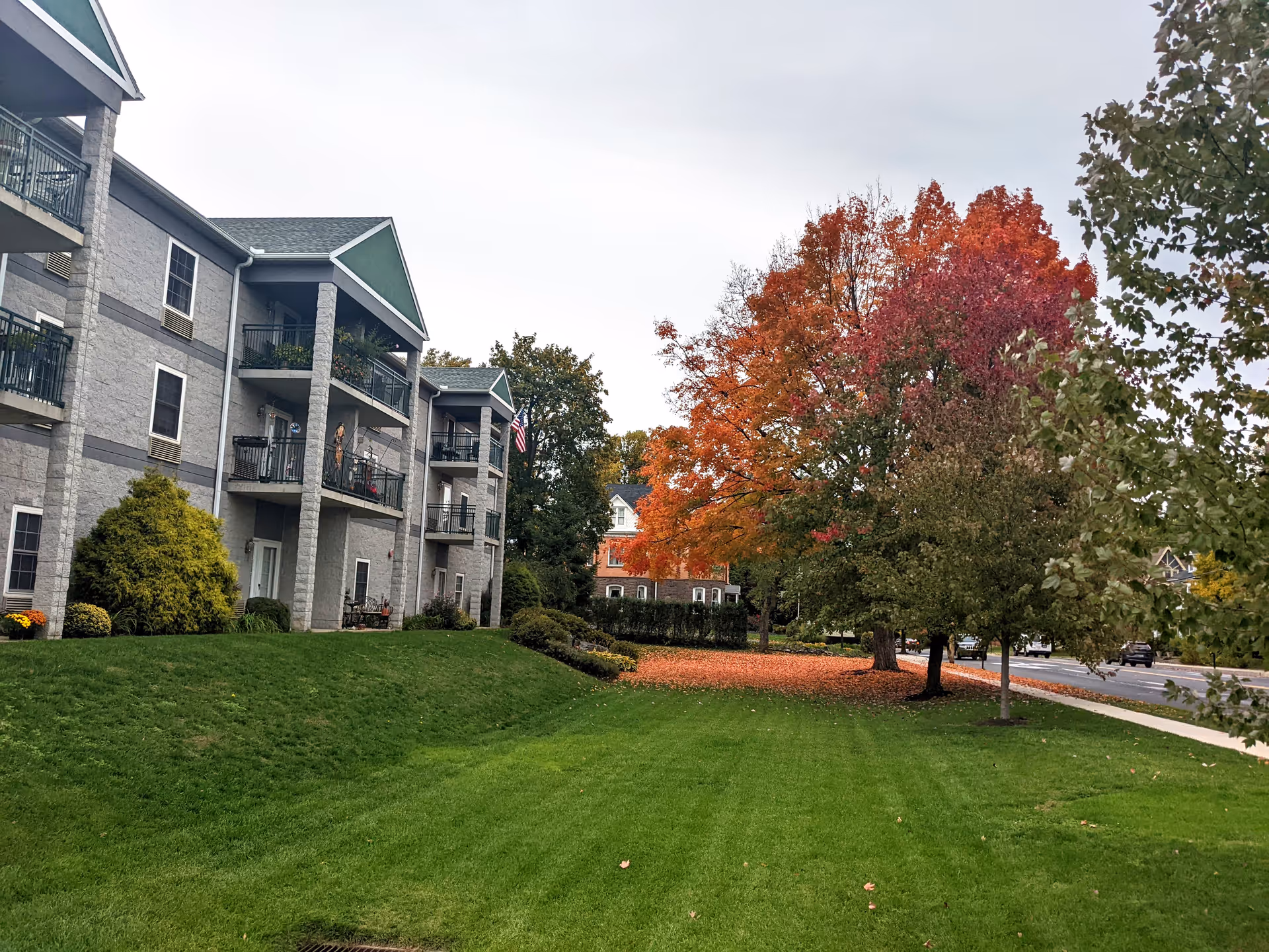 Exterior view of a multi-story senior living facility with balconies, green lawn, and trees with autumn foliage.