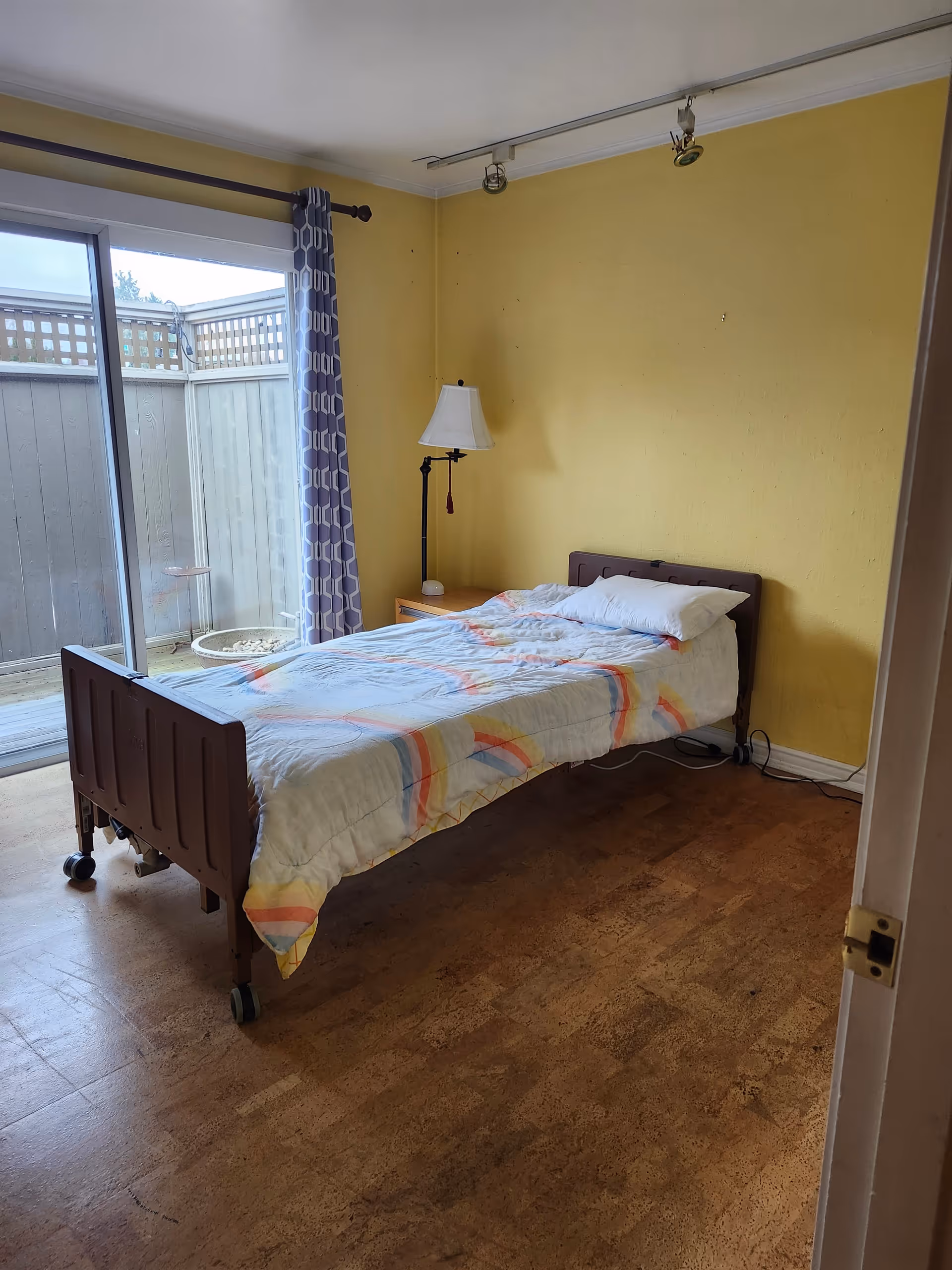 A simple bedroom with a single bed on wheels covered with a white blanket with colorful stripes. There is a wooden nightstand with a lamp next to the bed. The walls are painted yellow and there is a large sliding glass door with a curtain leading to an outdoor fenced area.