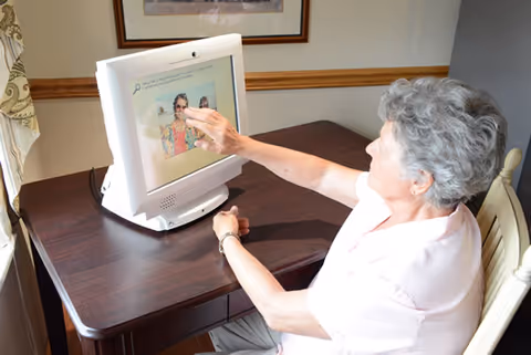 An elderly woman with short gray hair sitting at a wooden table, interacting with a touchscreen computer displaying a photo of a woman in sunglasses. The room has beige walls with a wooden chair rail and a framed picture hanging above the table.