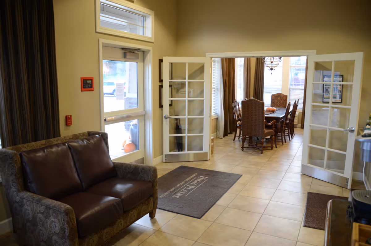 Interior view of a senior living facility showing a small seating area with a patterned armchair and brown leather cushions near a glass door entrance. Beyond the entrance, there is a room with a dining table surrounded by chairs, large windows with curtains, and a chandelier overhead. The floor is tiled and there are mats near the doors.