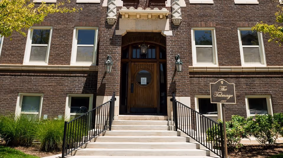 Front entrance of a brick building with a set of stairs leading up to a wooden door with a circular window. There are black metal railings on both sides of the stairs, two wall-mounted lanterns flanking the door, and a sign on the right side that reads 'The Talmage 1927'. Green shrubs and grass are visible around the entrance.