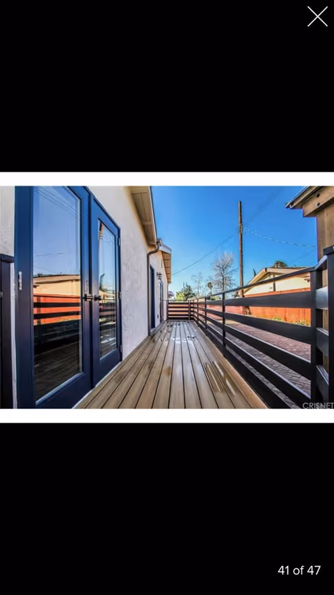 A narrow wooden deck or balcony attached to a white building with blue-framed glass doors on the left side. The deck has a black railing on the right side and overlooks a paved area with some trees and utility poles in the background under a clear blue sky.