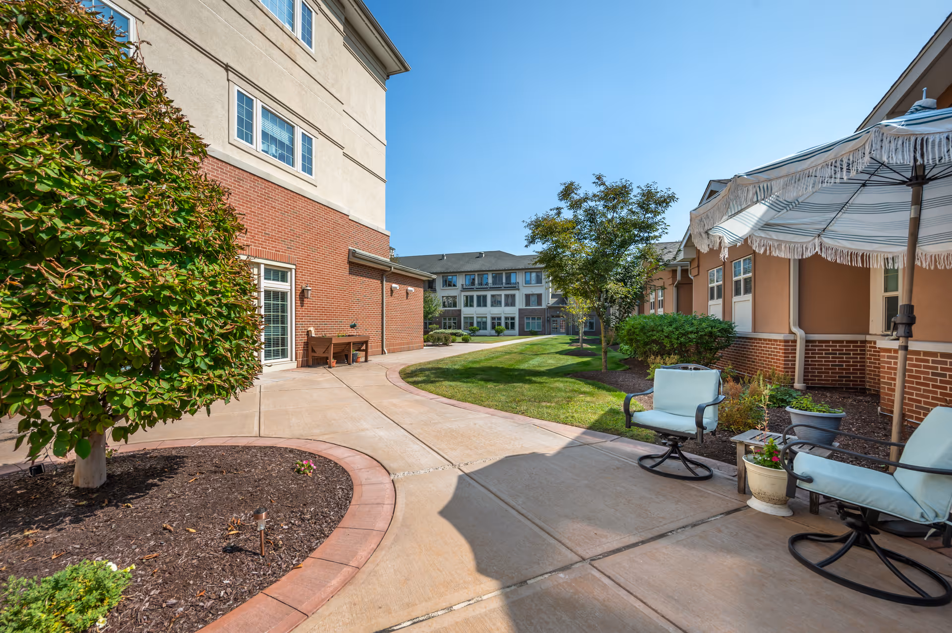 Sunny courtyard of a senior living facility with brick buildings, paved walkways, patio chairs and an umbrella, and landscaped lawn.