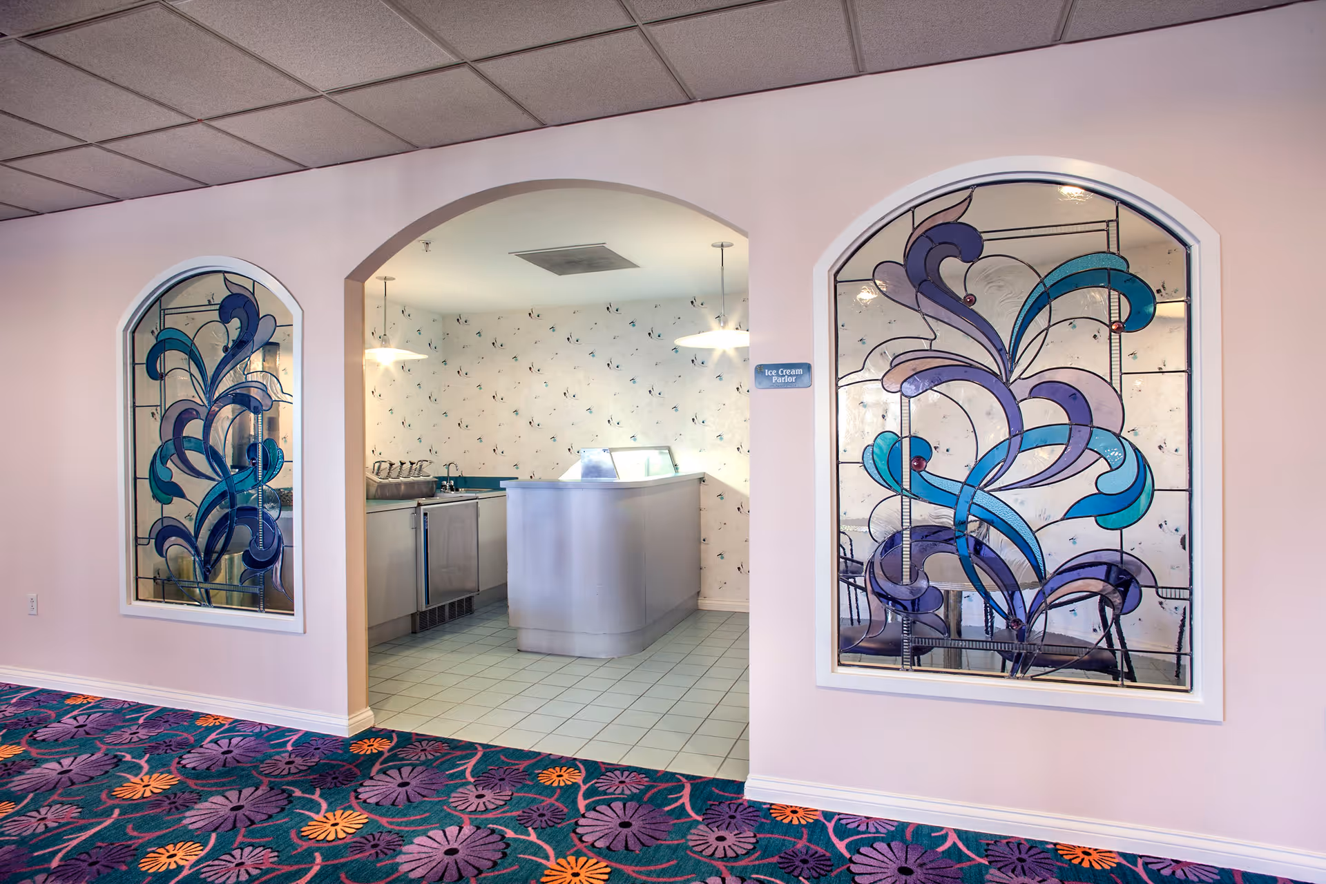 Interior view of an ice cream parlor area in a senior living facility with a counter and stainless steel appliances. The walls are light pink with two large decorative stained glass windows featuring blue and purple abstract designs. The floor has a colorful carpet with purple and orange floral patterns.
