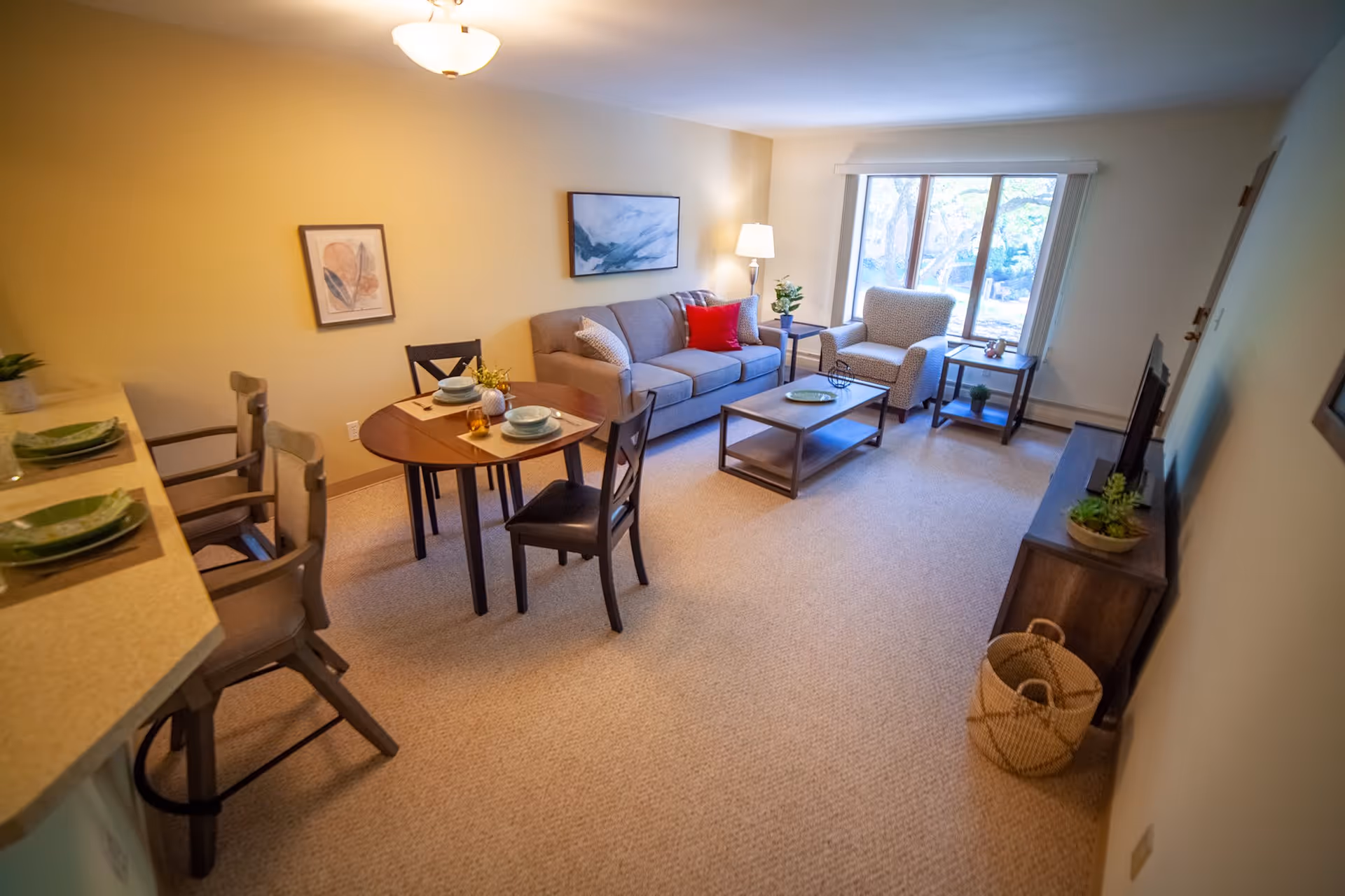 A cozy living room with a beige sofa adorned with pillows, including a red one, and a patterned armchair next to a window with a view of trees outside. A wooden coffee table and side tables with plants and decorative items are present. A small round dining table set for two is near the sofa, and two bar stools are positioned at a kitchen counter. The room has beige carpet and light-colored walls with framed artwork.
