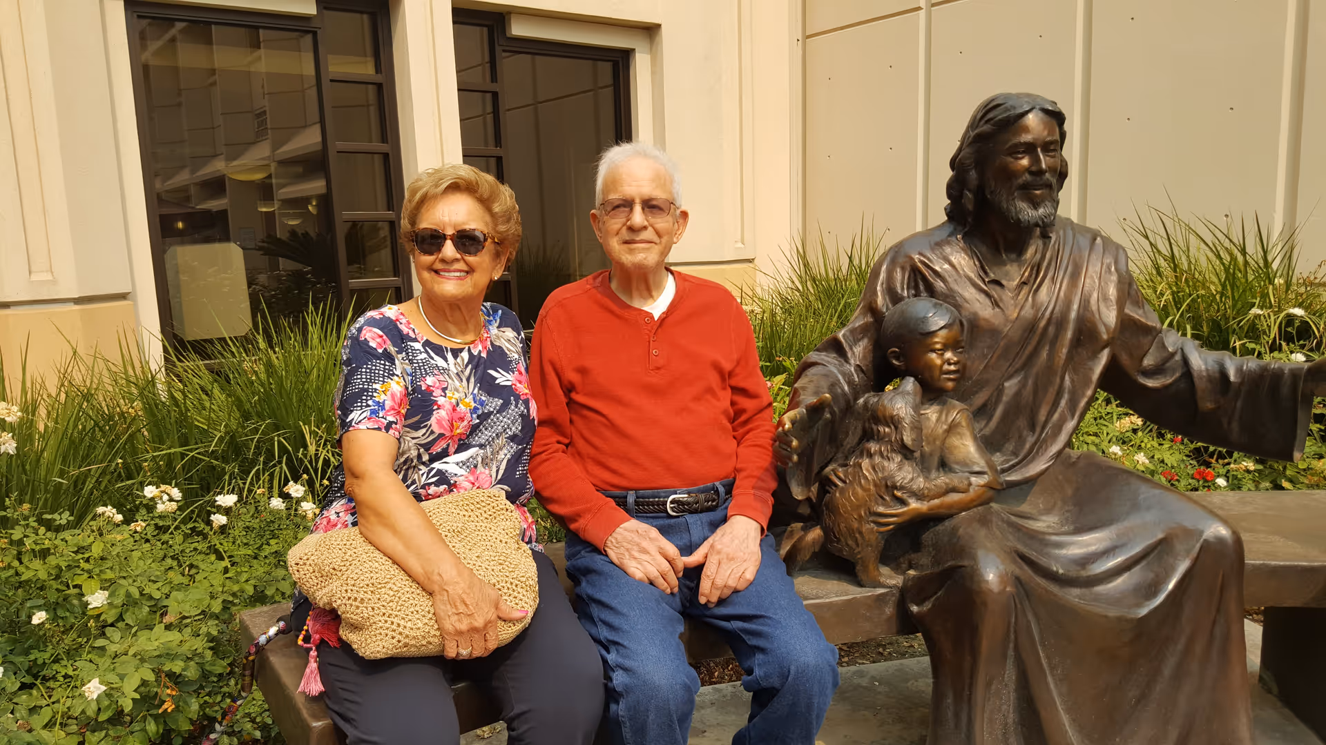 An elderly man and woman sitting on a bench outdoors next to a bronze statue of a man with a child and a dog. The woman is wearing sunglasses, a floral shirt, and holding a woven bag. The man is wearing glasses, a red long-sleeve shirt, and blue jeans. Behind them are green plants and a building with windows.