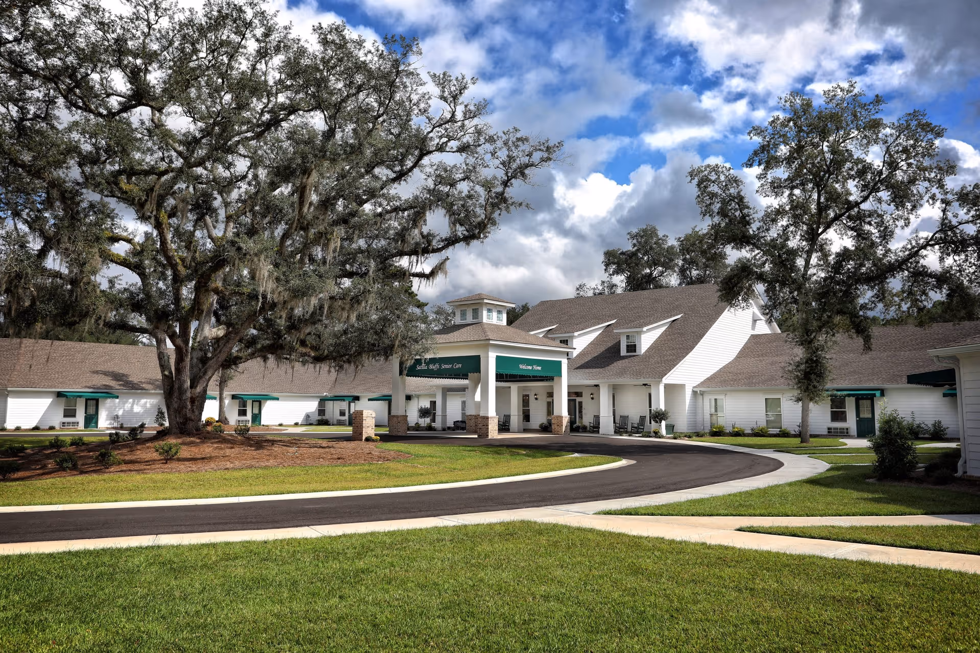 Exterior view of Satilla Bluffs Senior Care building with white siding, green awnings, and a covered entrance. Large trees and well-maintained grass surround the building under a partly cloudy sky.