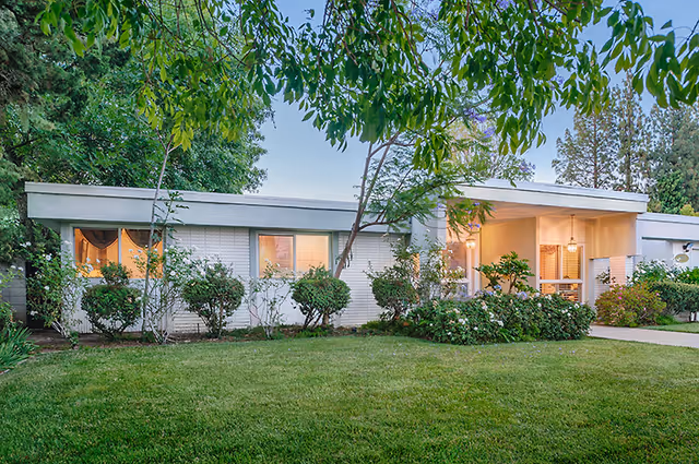 Single-story building with a flat roof surrounded by green bushes and trees, with a well-maintained lawn in front. The building has several windows, some with lights on inside, and a covered entrance area with outdoor lighting.