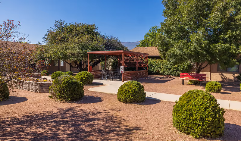 Outdoor area at Verde Valley Assisted Living featuring a paved walkway, neatly trimmed bushes, a red bench, and a shaded pergola with outdoor seating. Trees and shrubs surround the area under a clear blue sky.
