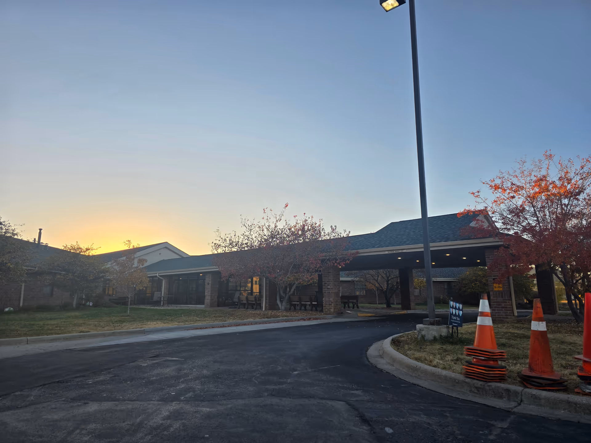 Exterior view of a single-story brick building with a covered entrance and a driveway, surrounded by trees with autumn foliage and traffic cones near the curb during sunset.