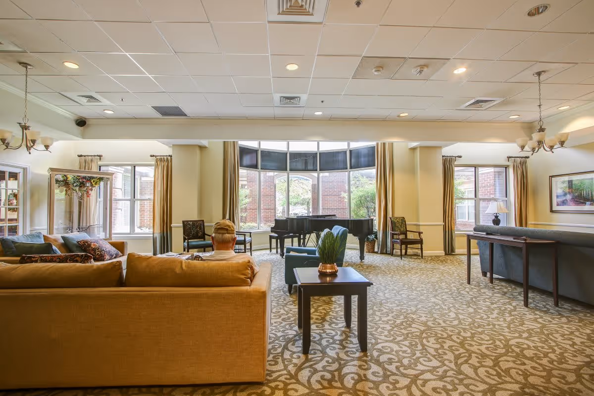A spacious and well-lit living room area with large windows allowing natural light to fill the space. The room features a grand piano near the windows, multiple chairs, and sofas arranged for socializing. A person wearing a cap is seated on a tan sofa facing away from the camera. The carpet has an intricate pattern, and the walls are decorated with framed artwork and curtains.