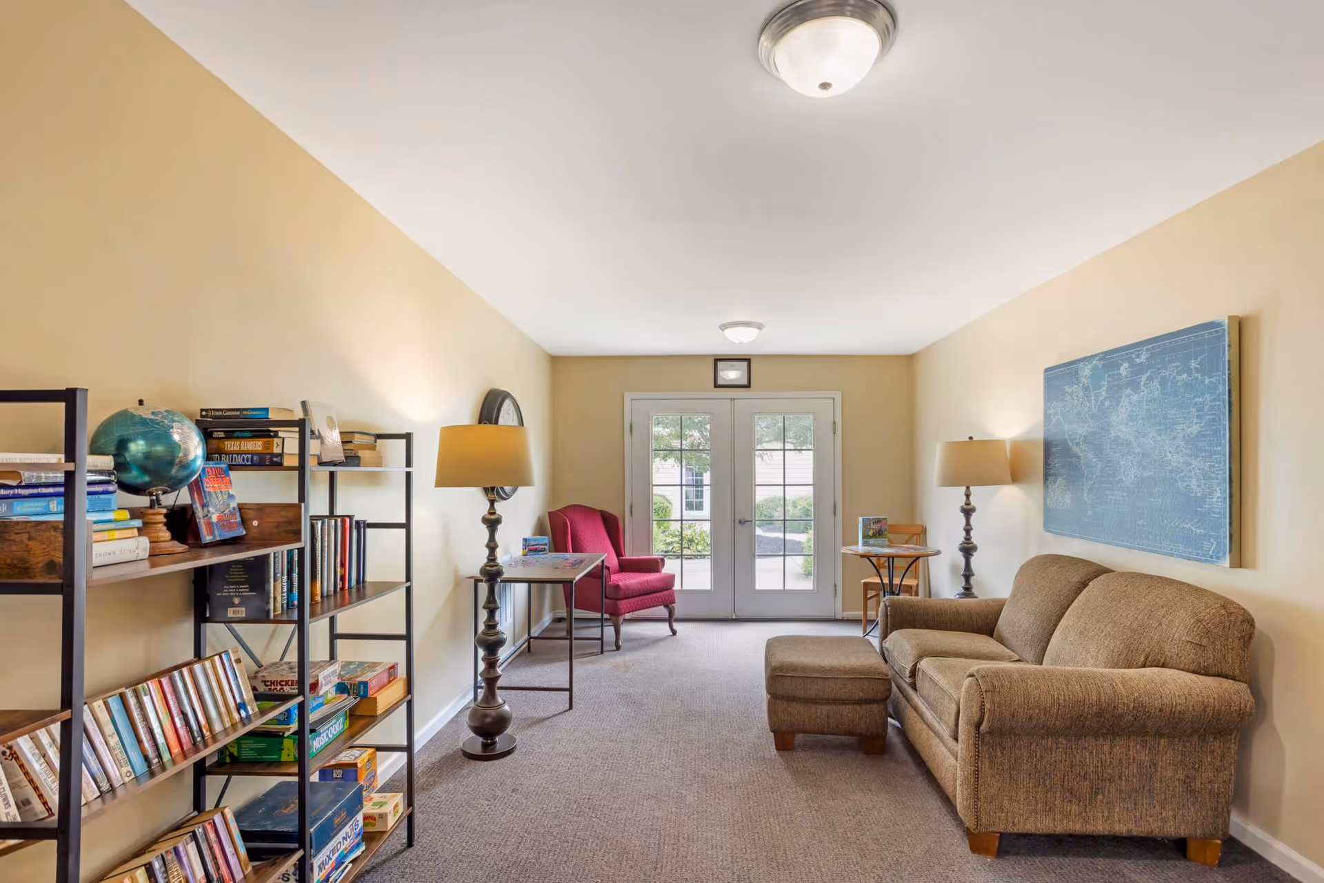 Cozy common room with a sofa and ottoman, bookshelves and lamps, a red armchair, and double glass doors leading outside.