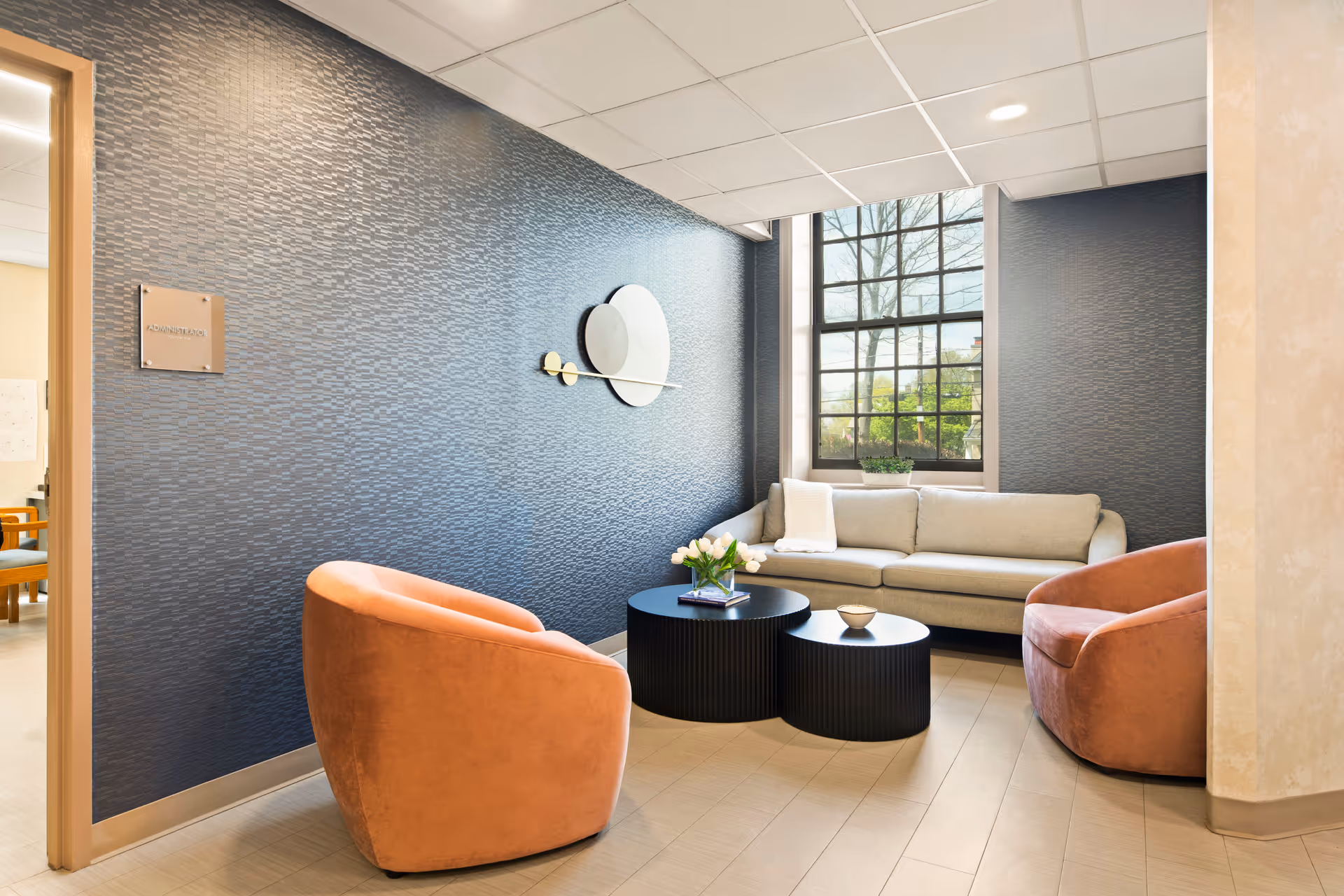A cozy seating area inside a facility with a beige sofa, two orange armchairs, and two black round coffee tables. The wall behind the sofa is textured and painted dark blue, featuring a modern circular wall decoration. A large window lets in natural light, and a small plant is placed on the windowsill. The floor is tiled, and there is a doorway leading to another room on the left.