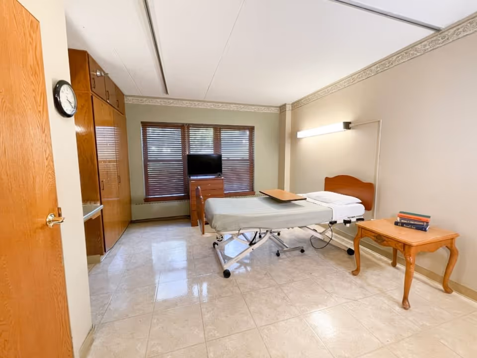 A senior living bedroom with an adjustable hospital-style bed, wooden furniture, a TV by the window with blinds, and tiled flooring.