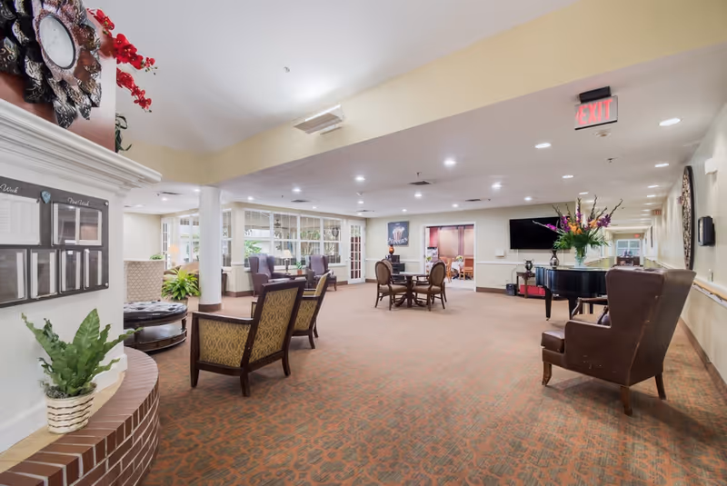 A spacious and well-lit common area in a senior living facility with patterned carpet, several upholstered chairs, a round table with chairs, a grand piano with a flower arrangement on top, and large windows letting in natural light. There is an exit sign on the ceiling and a decorative clock on the wall near a bulletin board.