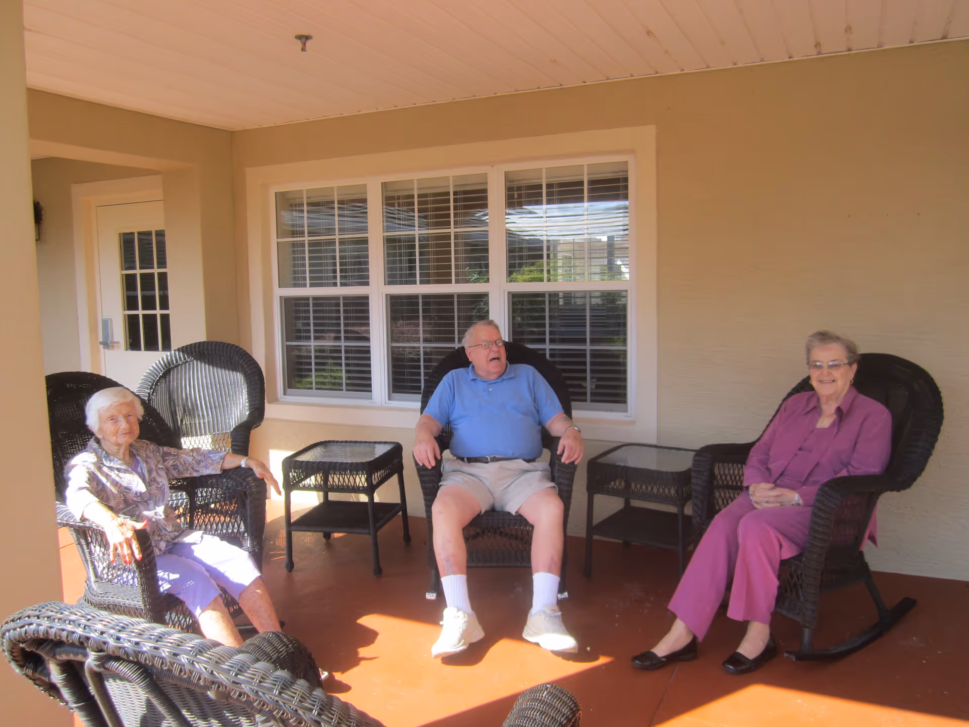 Three people sitting on wicker chairs on a covered porch in front of a window.