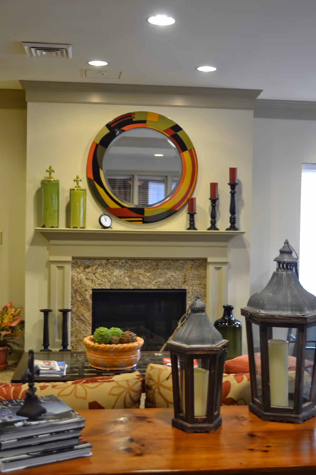 A cozy living room area featuring a fireplace with a granite surround. Above the fireplace is a colorful round mirror and decorative items including green jars, a small clock, and black candle holders with red candles. In front of the fireplace is a glass coffee table with a bowl of decorative balls. The foreground shows a wooden table with two large lantern-style candle holders and a stack of books. The room has warm, inviting decor with patterned cushions on the sofa.