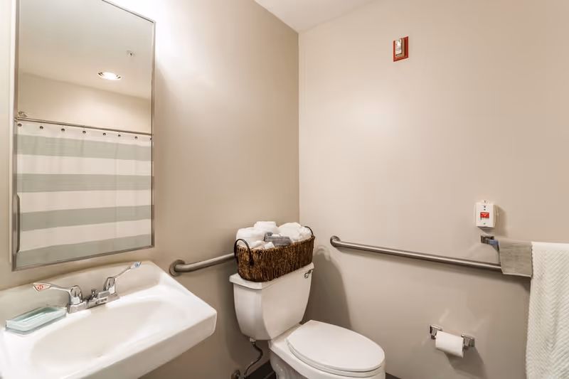 Accessible bathroom with a sink and mirror, a toilet topped with a basket of towels, grab bars, and a striped shower curtain.
