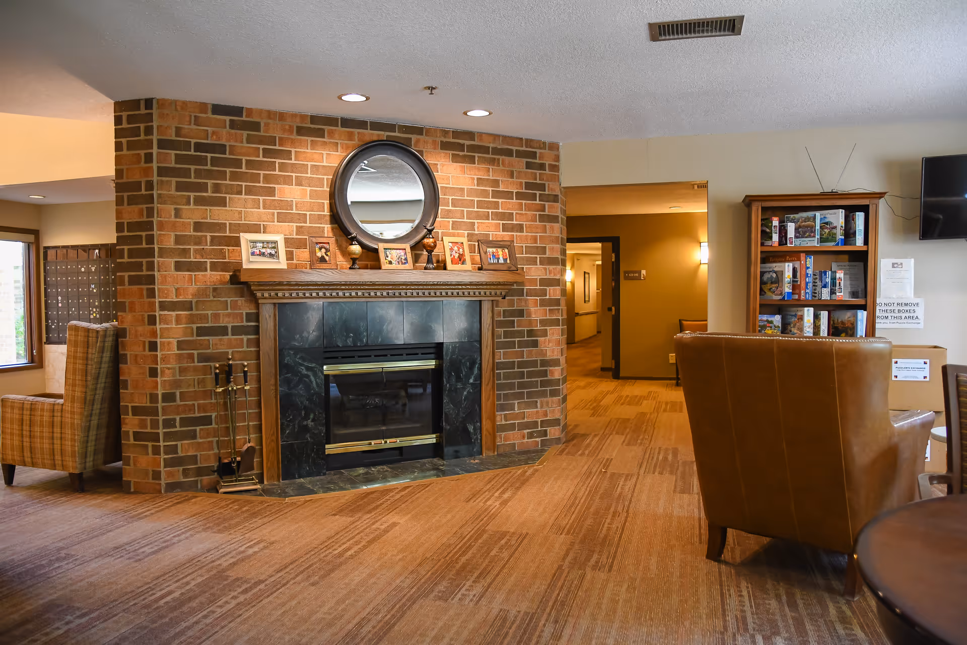 A cozy common area with a brick fireplace featuring a round mirror and framed photos on the mantel. The room has carpeted flooring, a brown leather chair, a wooden bookshelf filled with books and games, and a hallway leading to other rooms. Warm lighting creates a welcoming atmosphere.