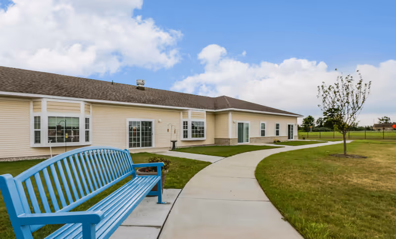 Outdoor view of a single-story beige building with multiple windows and sliding glass doors, a curved concrete walkway, a blue bench, a small tree, and a grassy lawn under a partly cloudy sky.