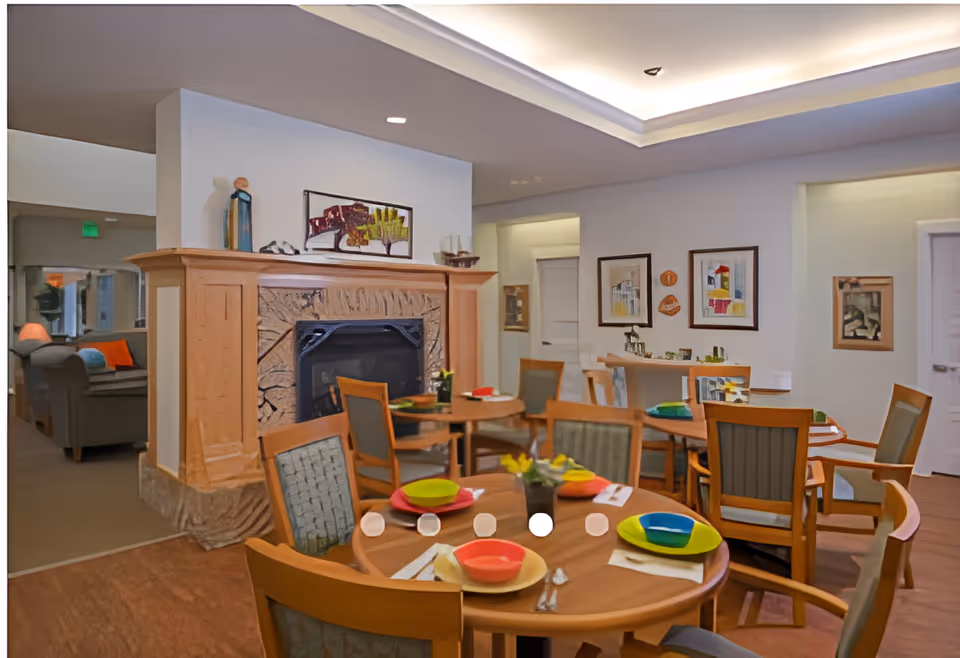 A cozy dining area in a senior living facility with round wooden tables set with colorful plates and bowls. The room features a stone fireplace with a wooden mantle, decorated with artwork and small decorative items. Several wooden chairs with cushioned seats surround the tables. The walls are adorned with framed pictures, and soft lighting creates a warm atmosphere.