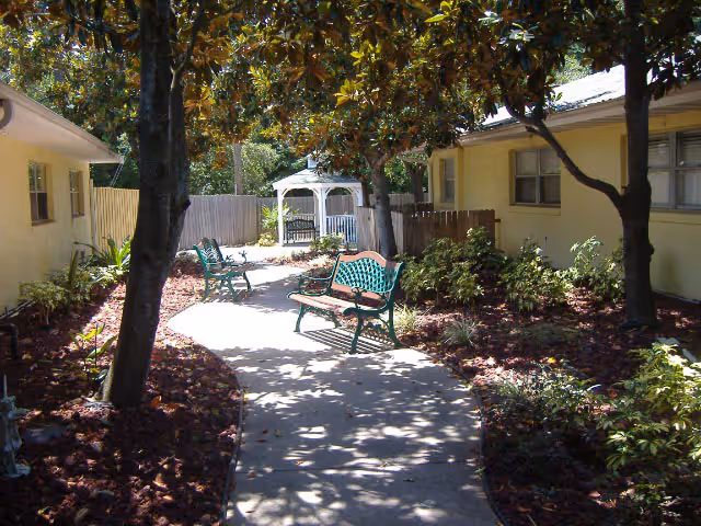 A shaded outdoor garden area with a curved concrete pathway, green benches, trees, and plants. There is a white gazebo at the end of the path, surrounded by two single-story yellow buildings.