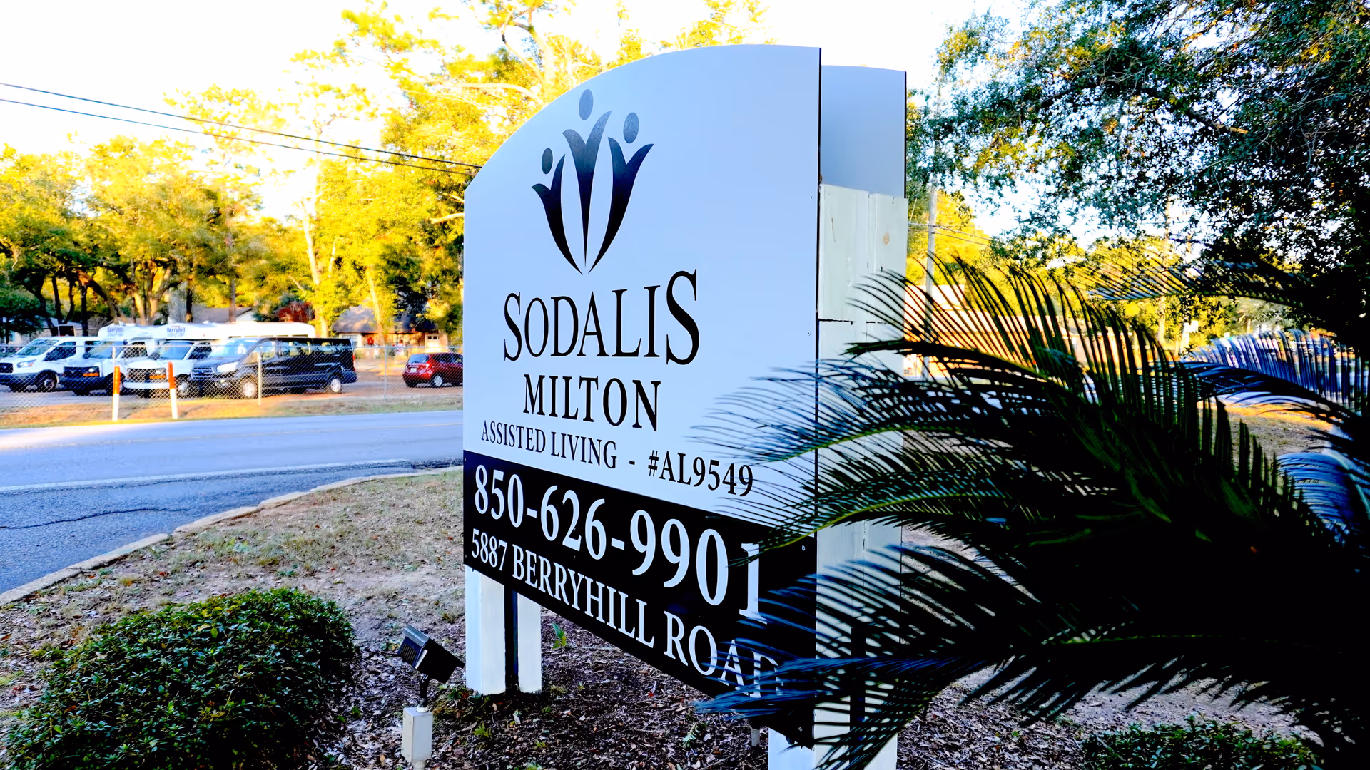 Outdoor view of a large white sign for Sodalis Milton Assisted Living located at 5887 Berryhill Road, with a phone number 850-626-9901. The sign is surrounded by greenery and trees, with a road and parked vehicles visible in the background.