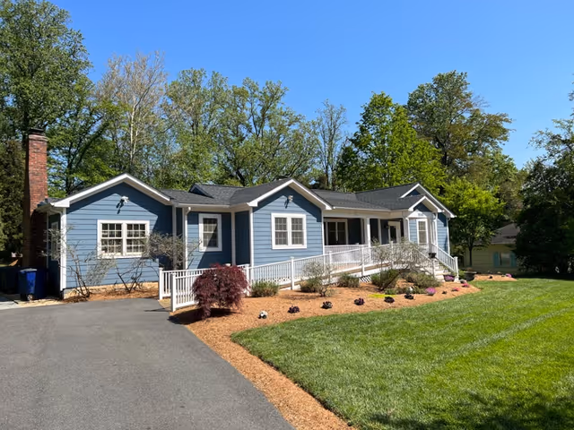 A single-story blue house with white trim and a black roof, featuring a wheelchair accessible ramp leading to the front porch. The house is surrounded by a well-maintained lawn, small shrubs, and trees in the background under a clear blue sky.