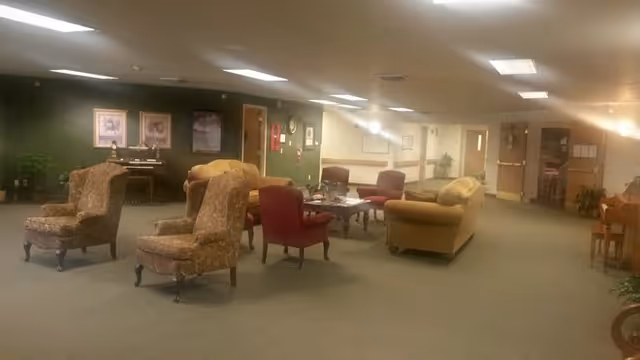 Open senior living common room with upholstered armchairs, sofas, coffee tables and a piano under fluorescent ceiling lights.