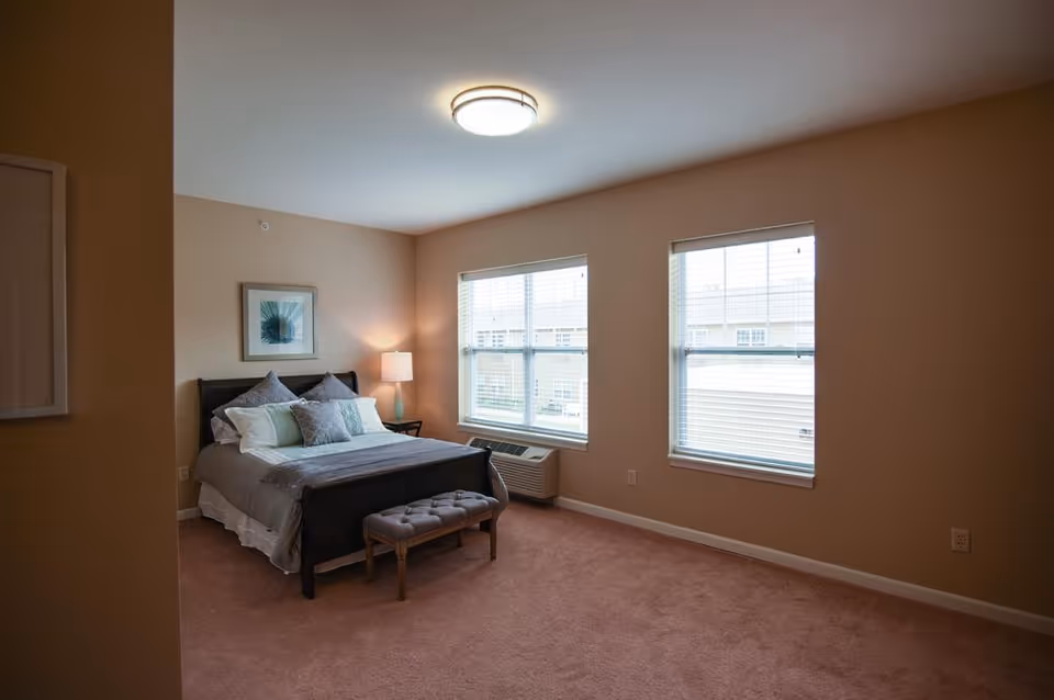 A bedroom with a neatly made bed featuring multiple pillows and a gray bedspread. There is a small cushioned bench at the foot of the bed, a bedside table with a lamp, and two large windows with blinds letting in natural light. The walls are painted beige and the carpet is light brown.