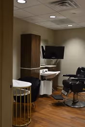 Interior view of a small grooming or barber room with a black barber chair, a wall-mounted TV, a white sink, a wooden cabinet, and a small round table with a black chair. The room has wood flooring and beige walls with recessed ceiling lights.